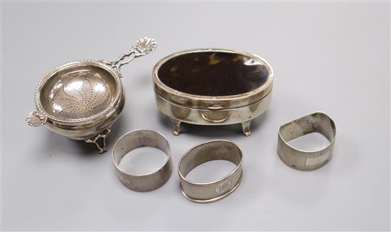 A George V silver trinket box, three serviette rings and a tea strainer and stand.